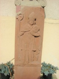 Le fût-stèle représente saint Nicolas, portant sur un livre les trois bourses dont il dota les jeunes filles.