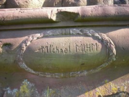 Le nom du sculpteur, Michael Mihm, est inscrit dans un cartouche entouré d'une guirlande végétale, situé sur la face du socle de la croix.