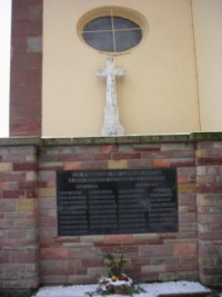 La croix est adossée à la façade de l'église, à gauche de l'entrée et derrière le monument aux morts communal.