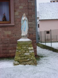Une statue de Notre-Dame de Lourdes est érigée dans le jardin d'une maison de la rue principale, sur un piédestal.
