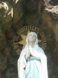 La statue de Notre-Dame de Lourdes est surmontée d'une auréole dorée, portant les paroles par lesquelles Elle s'est présentée à sainte Bernadette à Lourdes. La statue de Notre-Dame de Lourdes est surmontée d'une auréole dorée, portant les paroles par lesquelles Elle s'est présentée à sainte Bernadette à Lourdes.